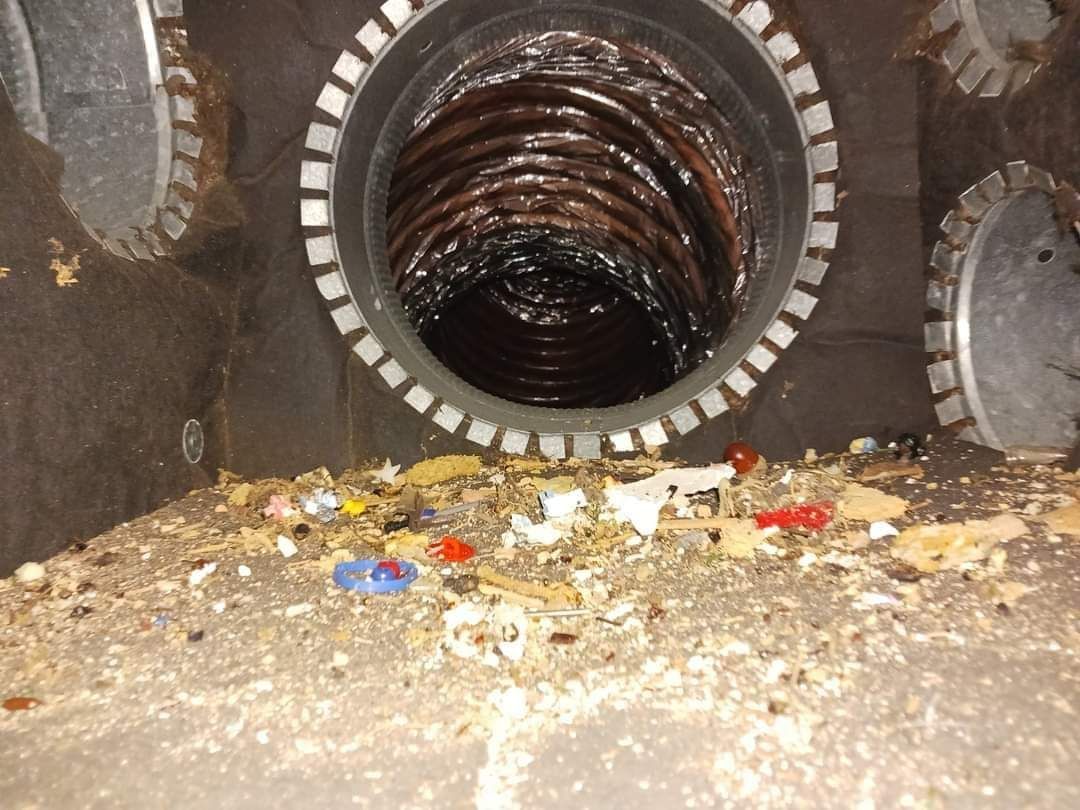 A rusty sewer pipe opening with debris scattered on the ground and trash near the entrance