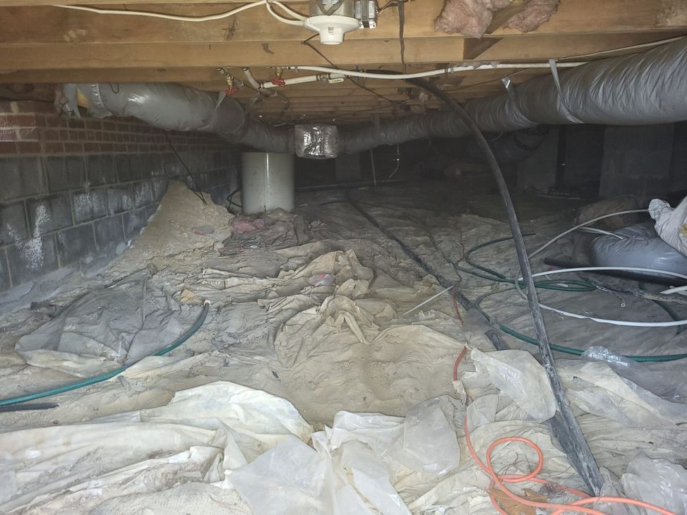 Crawlspace under a house with exposed insulation, ducting, wires, and debris on the ground.