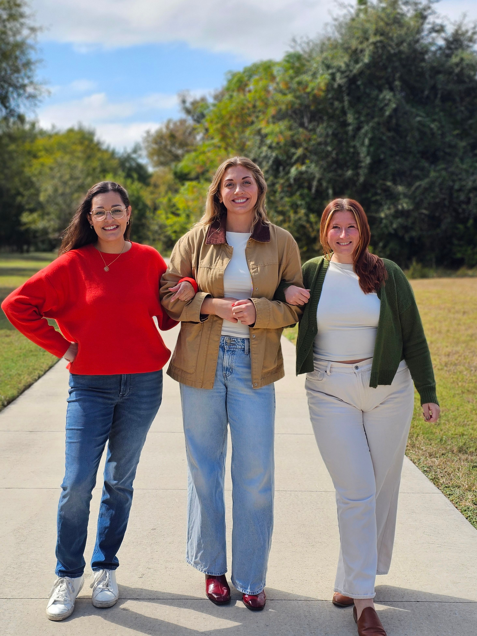 Three people walking on a sunny park path, smiling and dressed in casual clothes.