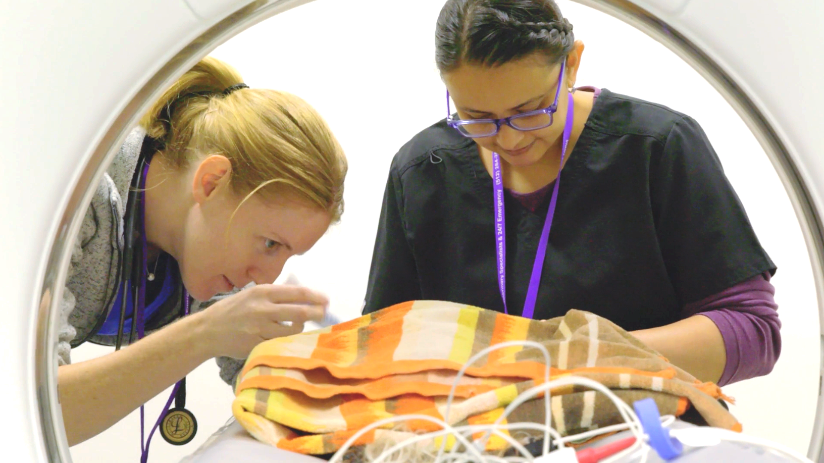 Two people lean over a swaddled baby in a clear incubator, examining it closely.