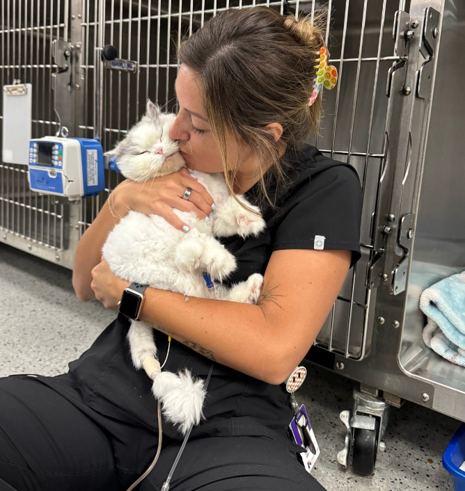 Woman kneels in a kennel, hugging a small white dog against stainless steel cages.