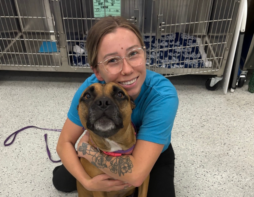 Woman in blue shirt hugging a brown dog in a kennel area, both looking at the camera.