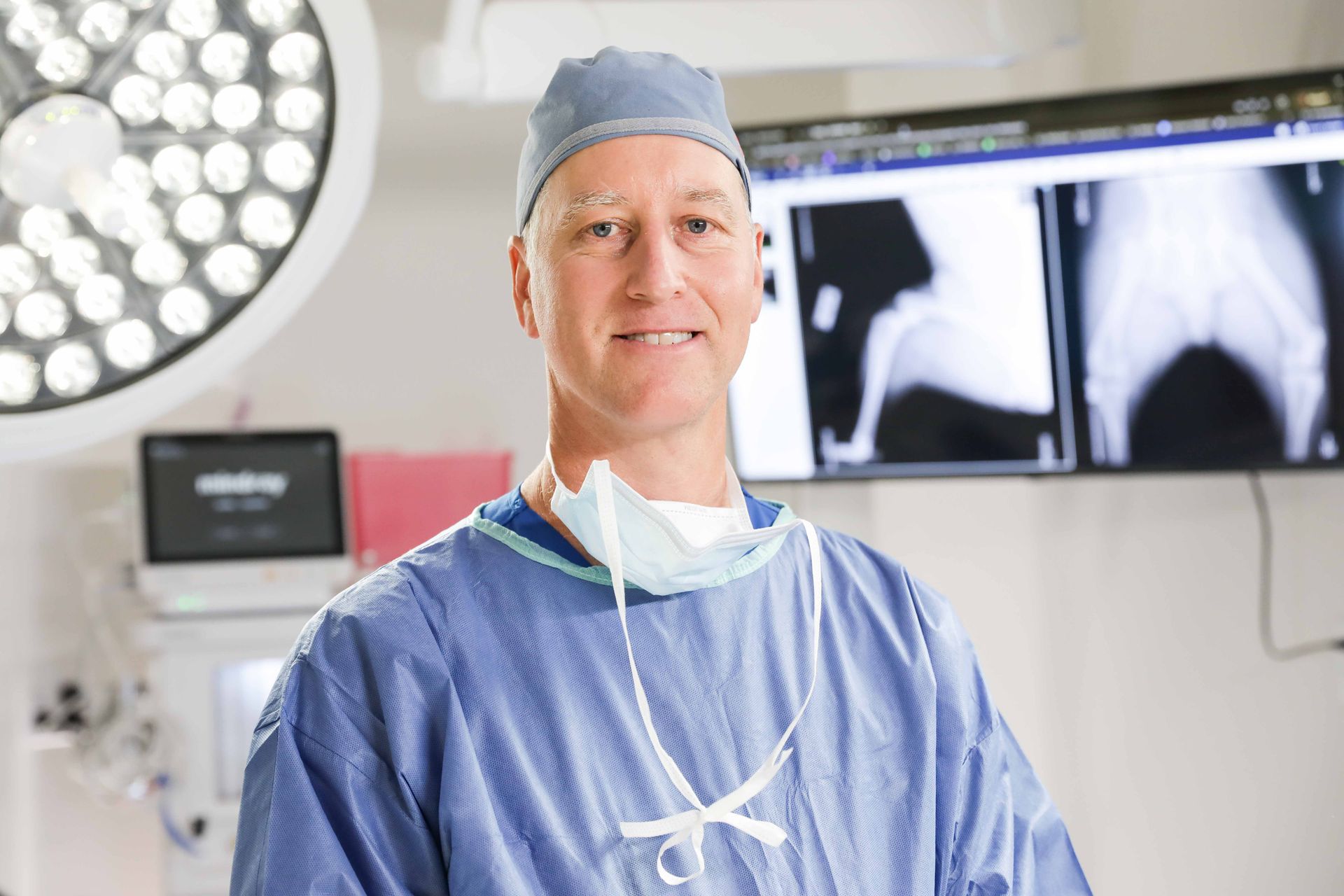 Surgeon in blue scrubs standing in an operating room beside X-ray monitors