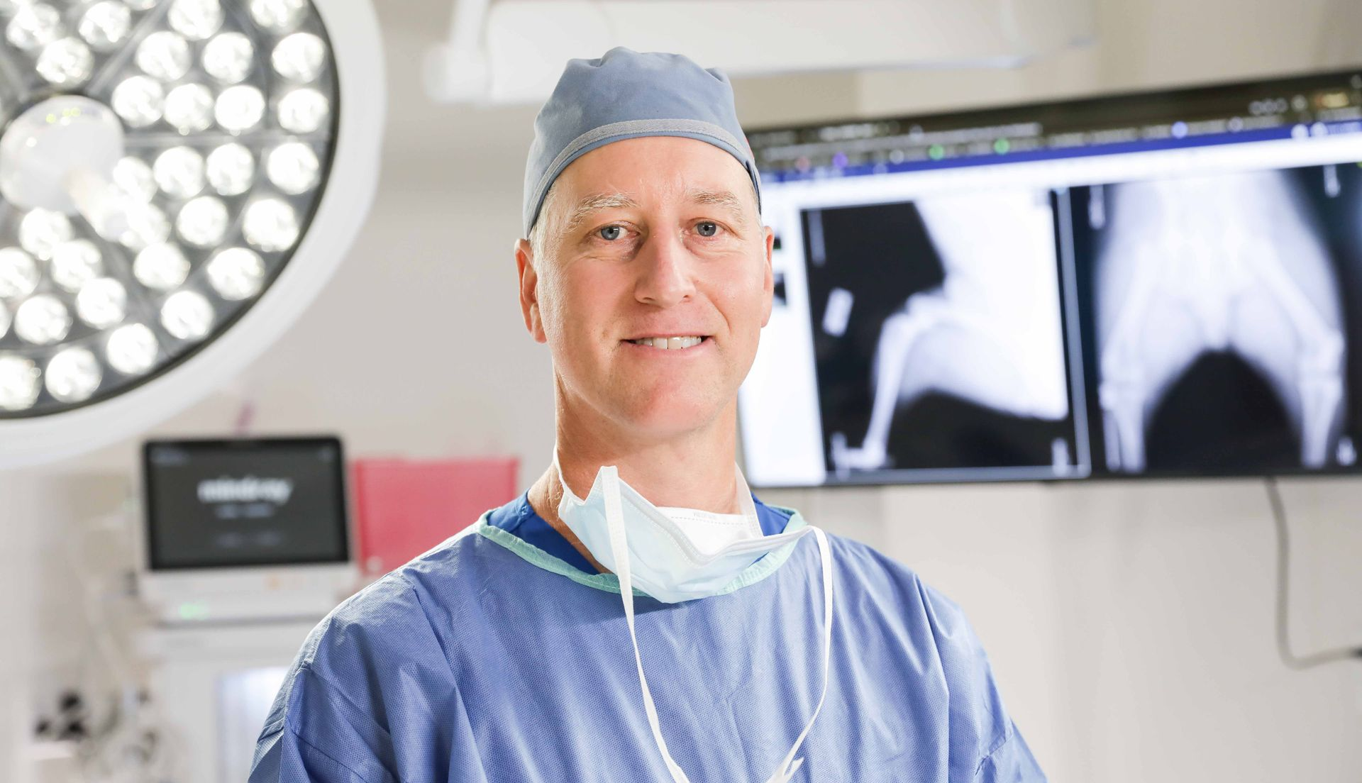 Surgeon in scrubs standing in an operating room with X-ray images on monitors behind him