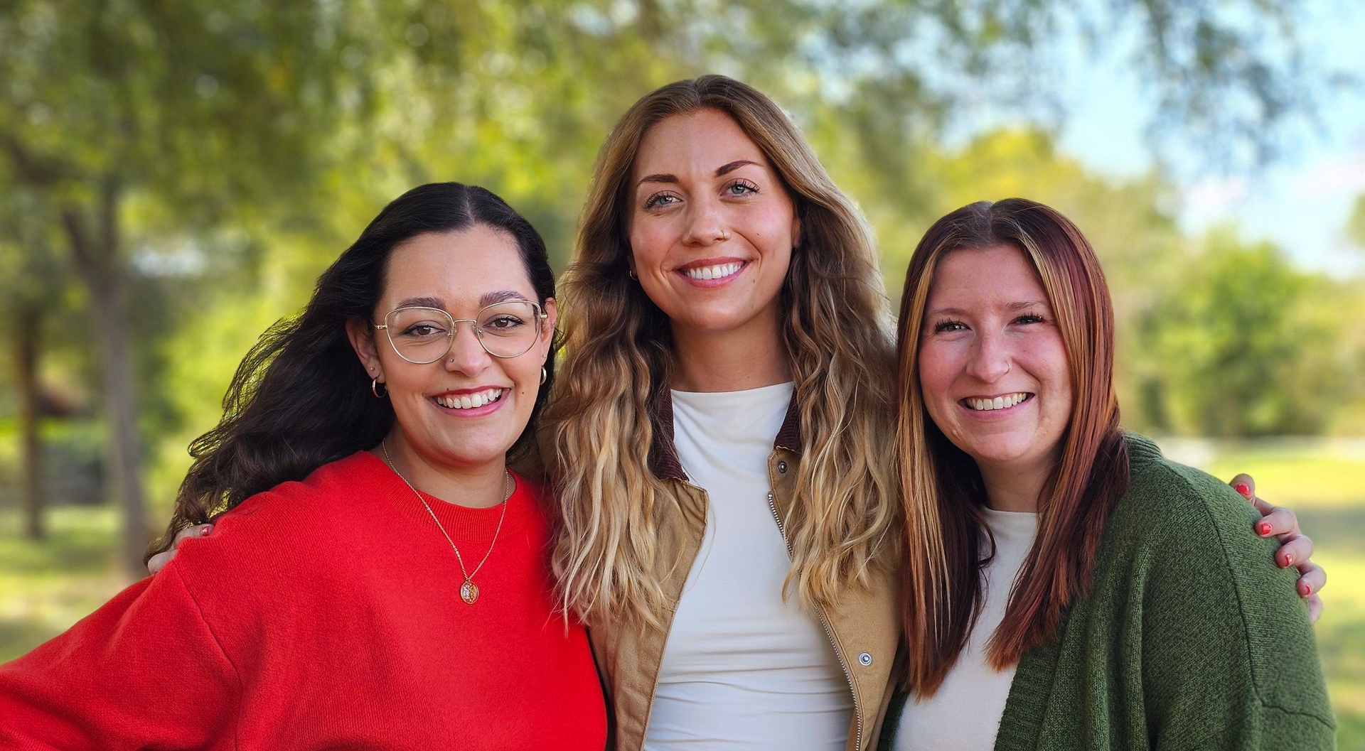 Three smiling people standing outdoors in colorful sweaters with trees in the background