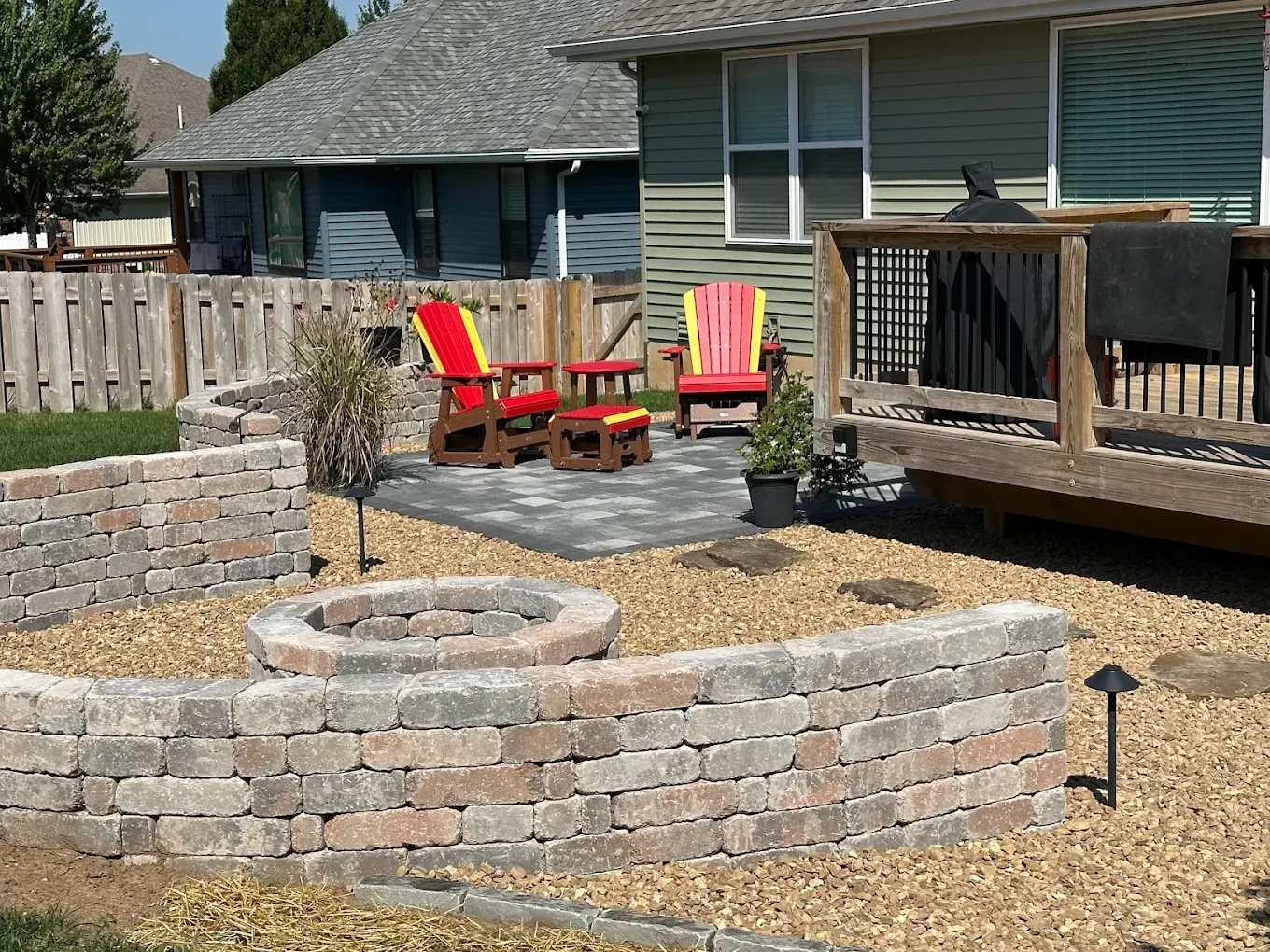 Backyard patio with stone fire pit, red-yellow chairs, and a house under a cloudy sky