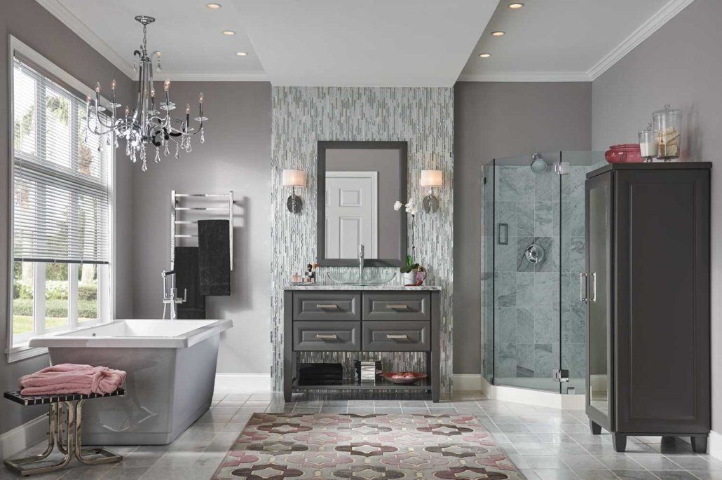 Elegant gray bathroom with chandelier, freestanding tub, vanity, glass shower, and patterned rug