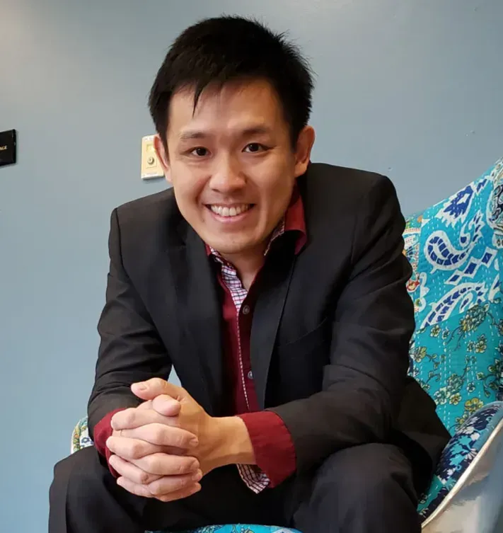 Smiling person in a black blazer seated indoors on a couch with blue patterned pillows.