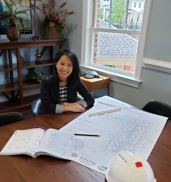 Woman smiling at a table with open plans and a hard hat in a bright office