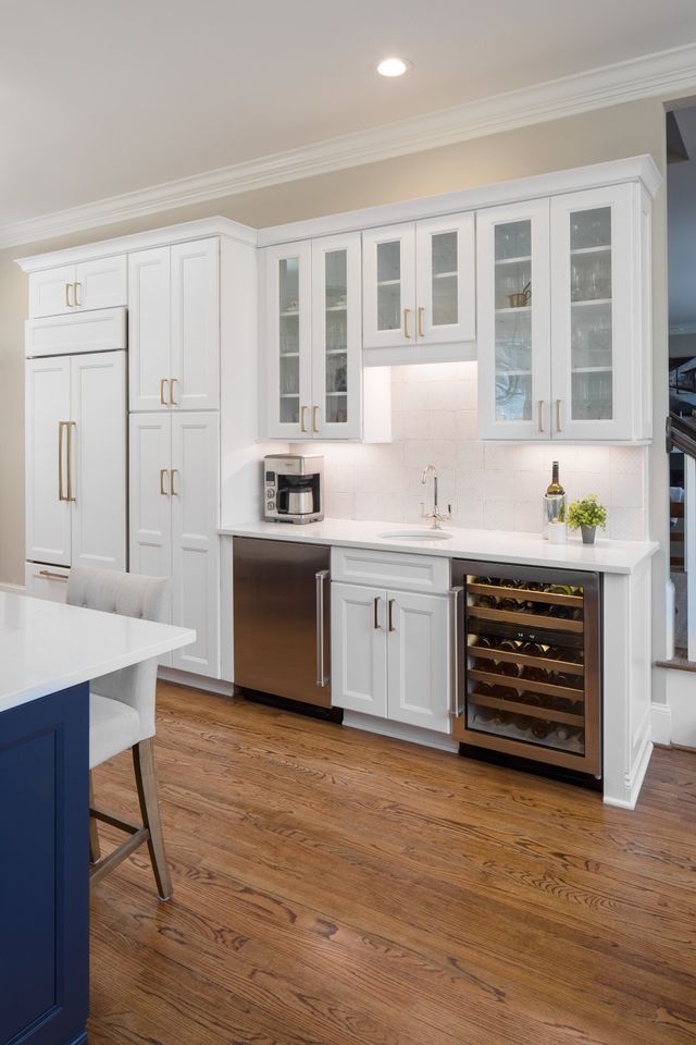 Modern white kitchen with glass-front cabinets, stainless fridge, wood floor, and dark blue island