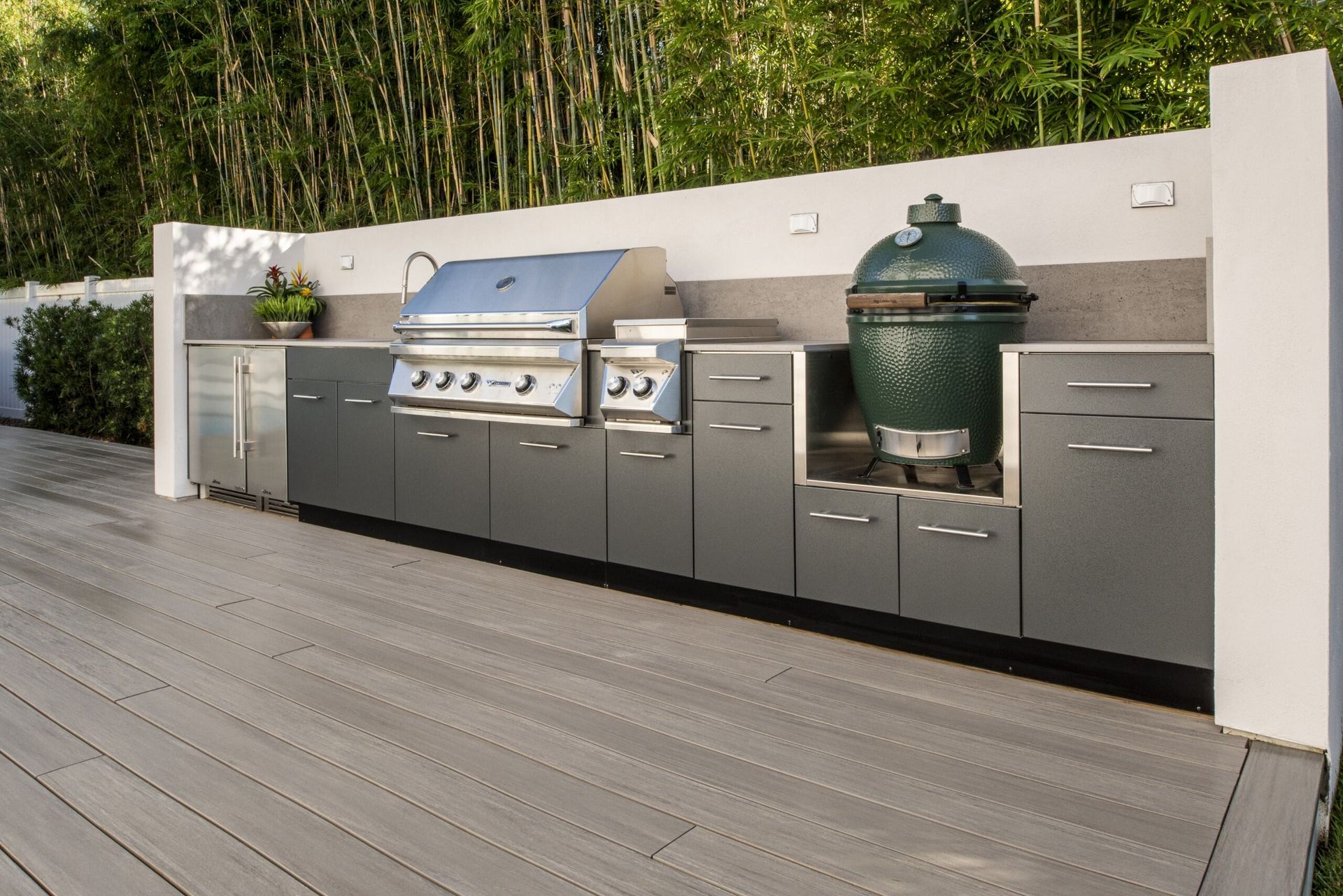 Outdoor stainless steel grill island with cabinets and a green ceramic smoker on a wooden deck