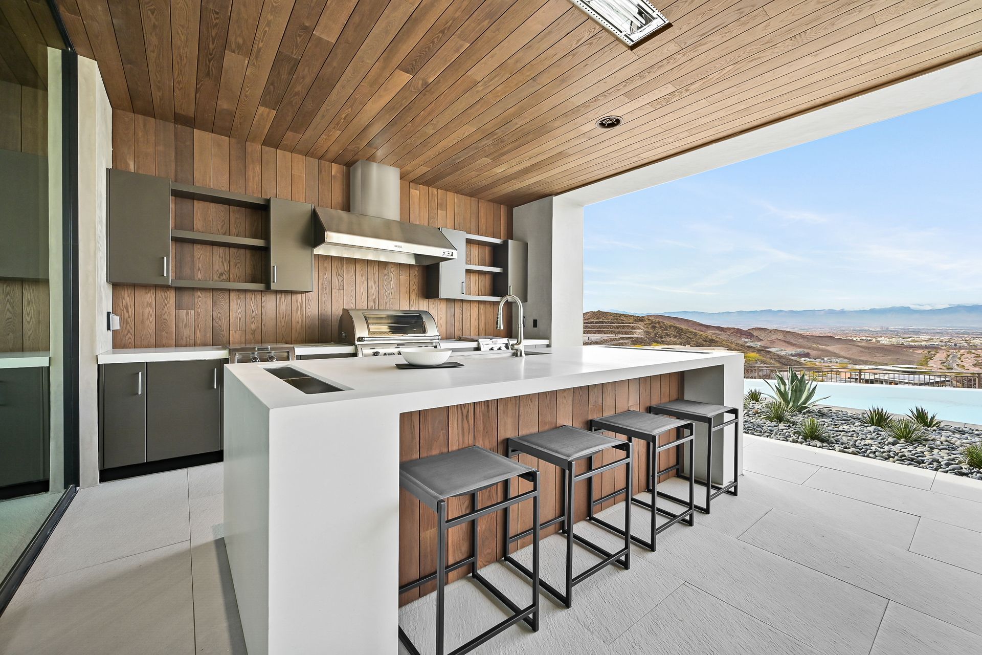 Modern outdoor kitchen with white island, bar stools, wood ceiling, and scenic lake view