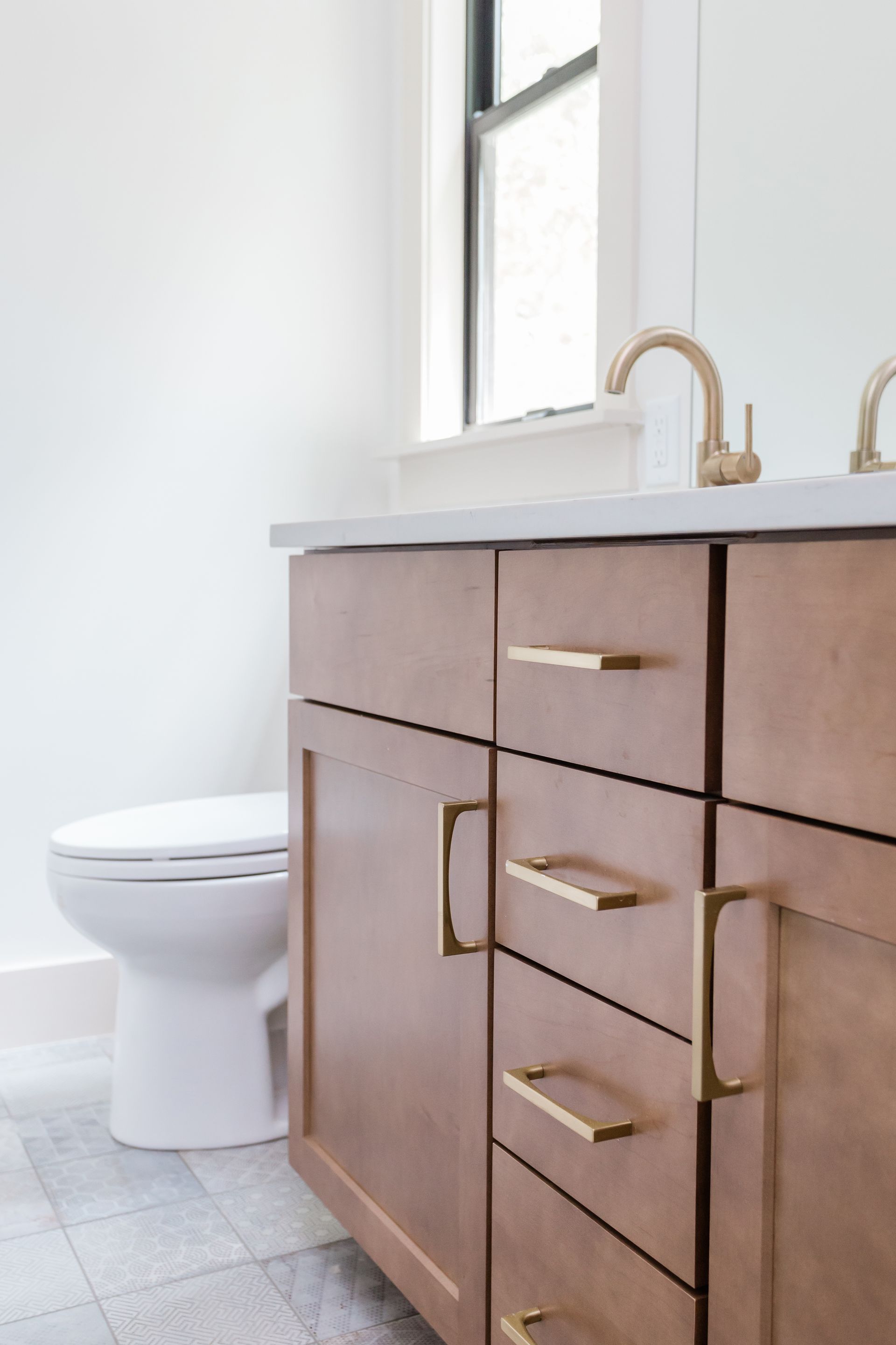 Modern bathroom vanity with wooden cabinets, gold faucet, and white toilet beside a window