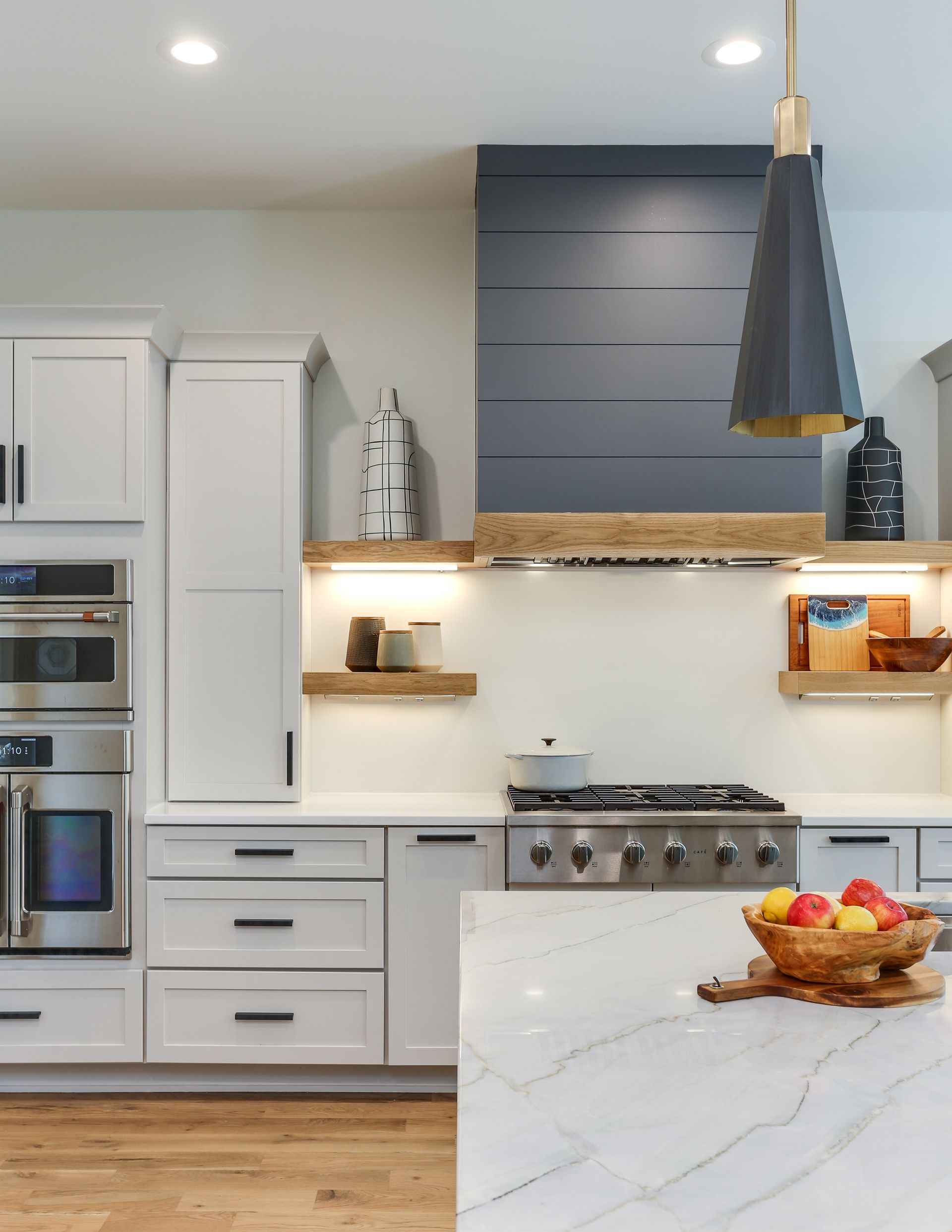 Modern white kitchen with gray range hood, wooden shelves, and marble island with fruit bowl.