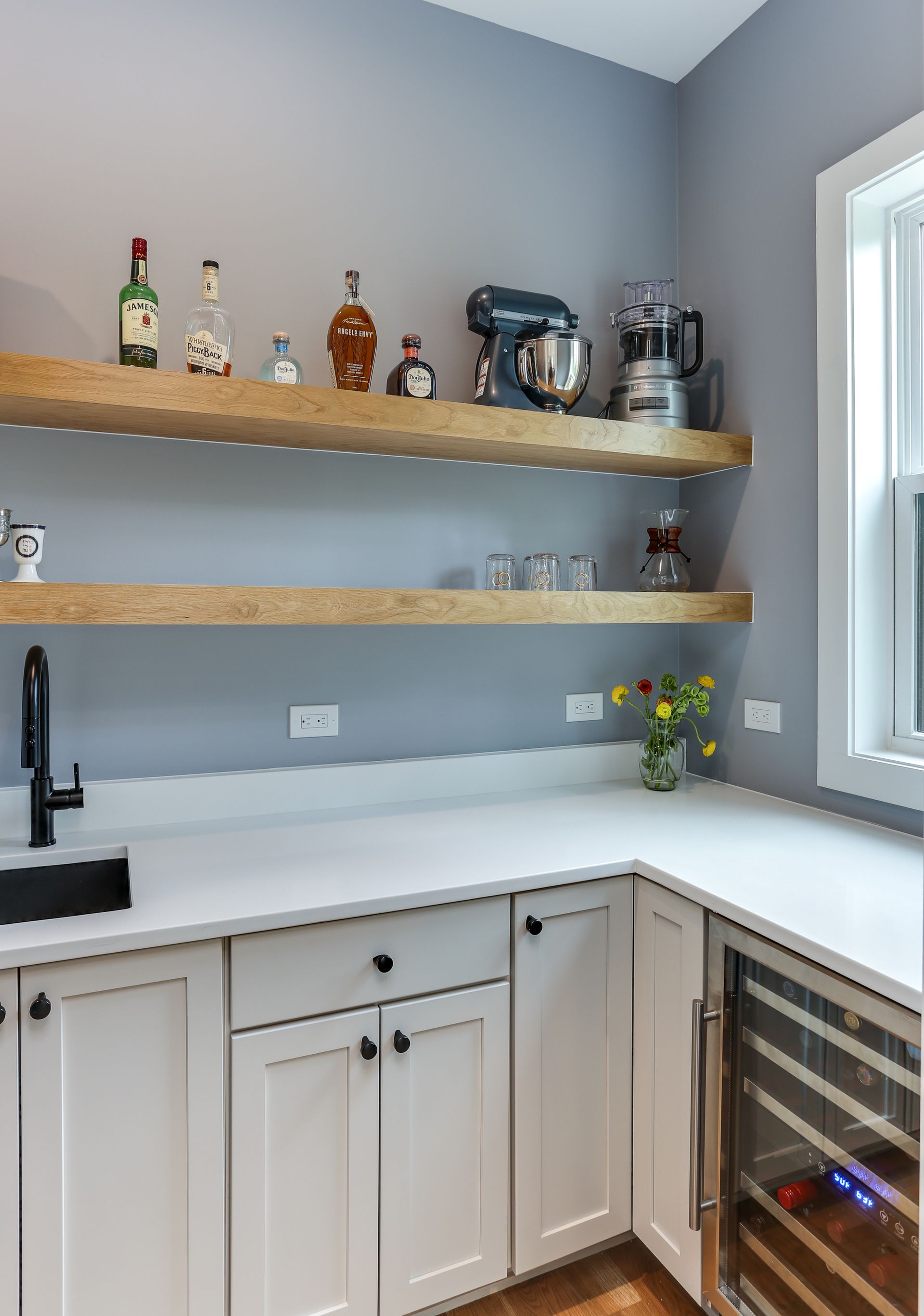 Modern home bar nook with floating shelves, white counter, black sink, and wine cooler by a window