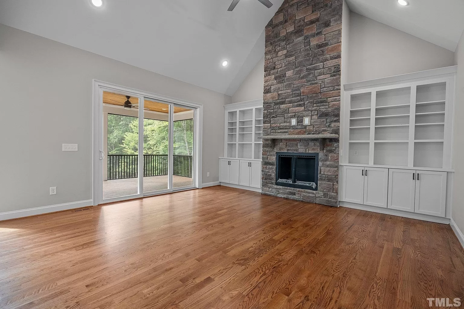 Bright living room with hardwood floors, vaulted ceiling, stone fireplace, built-in shelves, and glass doors to deck