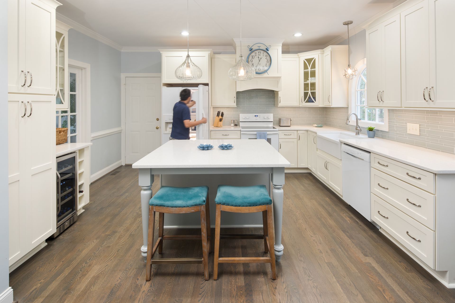 Bright white kitchen with a center island and two stools, and two people chatting near the back counter.