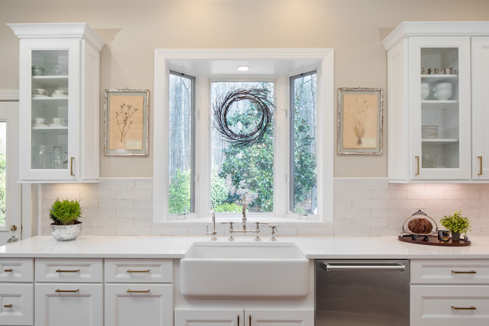 Bright white kitchen with farmhouse sink, marble counters, and a bay window with wreaths and greenery.