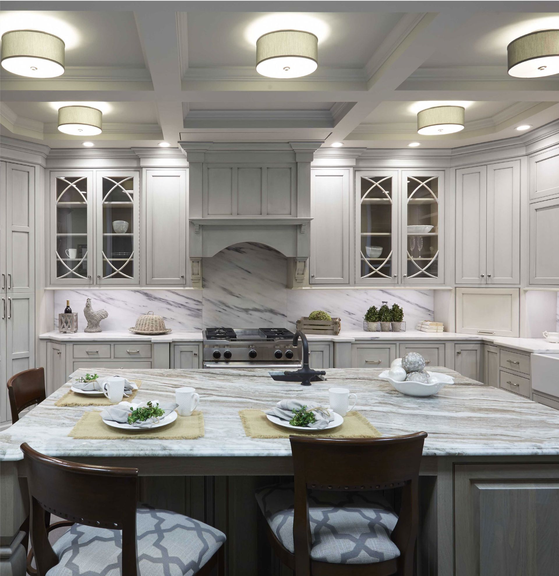 Elegant white kitchen with marble island, pendant lights, glass cabinets, and modern stove under a decorative hood