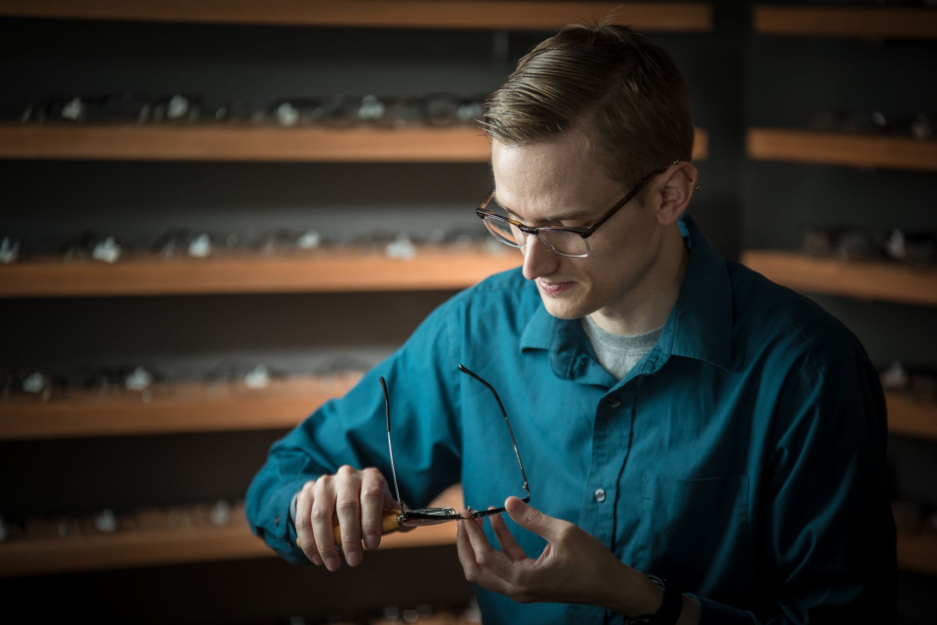 A man in a teal shirt repairs eyeglasses in a shop with a wall of frames.