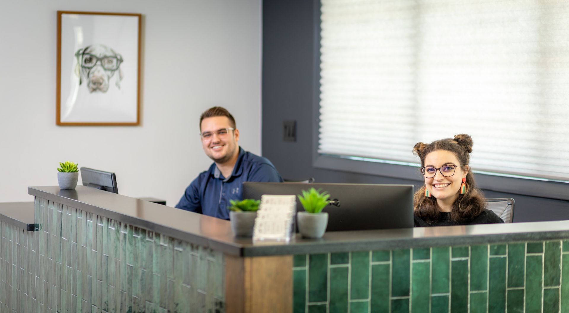 Two people smiling behind a reception desk. A green tile-fronted counter with a framed dog portrait hangs on the wall.