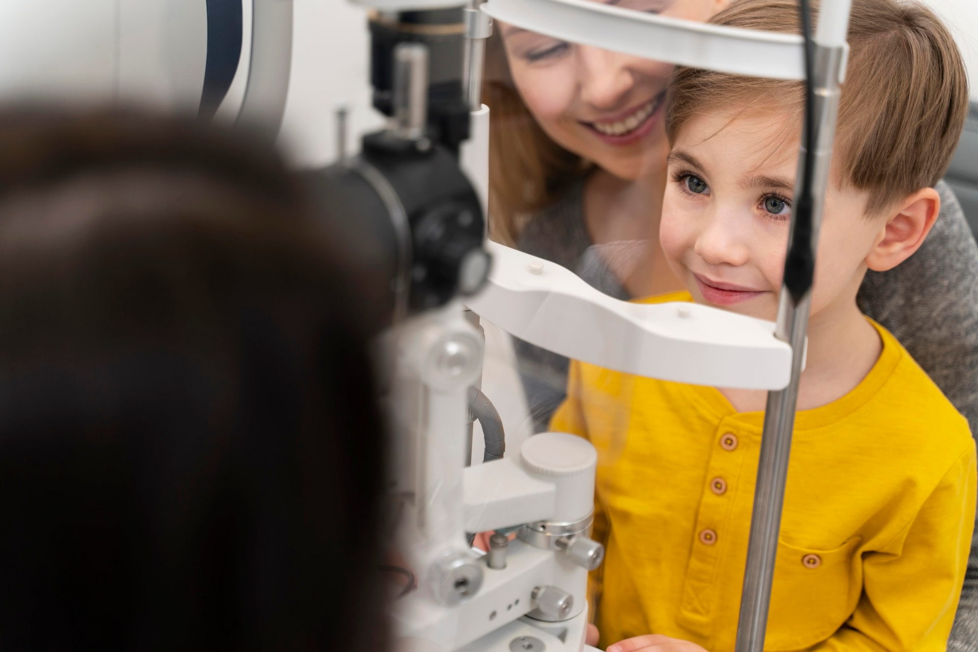 A young boy gets an eye exam with his mother present. He is looking into the machine and smiling, with the mother behind him.