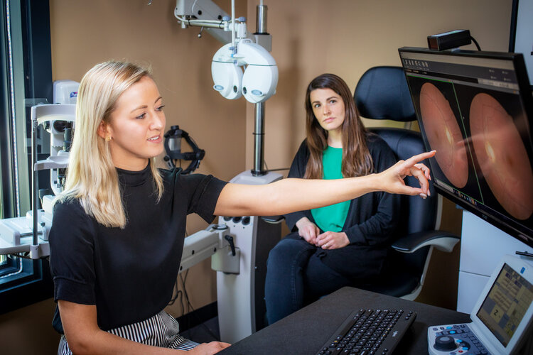 An optometrist points at a retinal scan displayed on a screen, explaining results to a seated patient in an examination room.