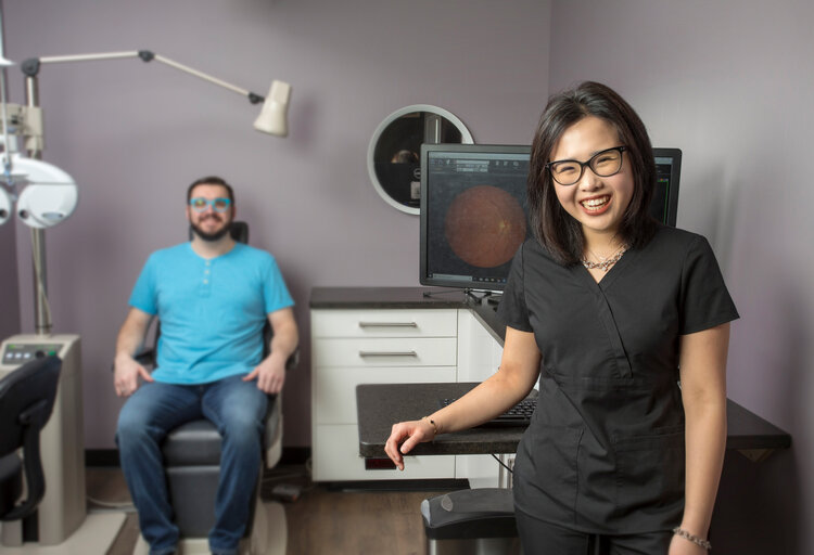 An Asian woman in glasses smiles at the camera in an eye doctor's office. A man sits in a chair, looking ahead.