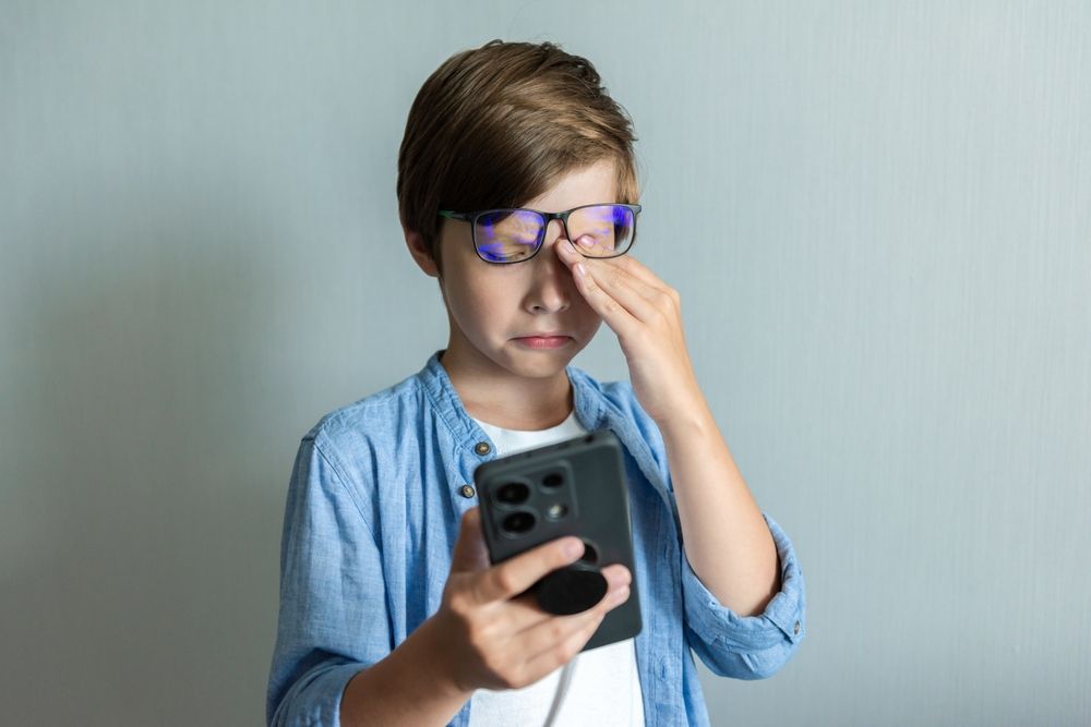 A young boy with glasses rubbing his eye while holding a smartphone, looking tired, against a light gray background.