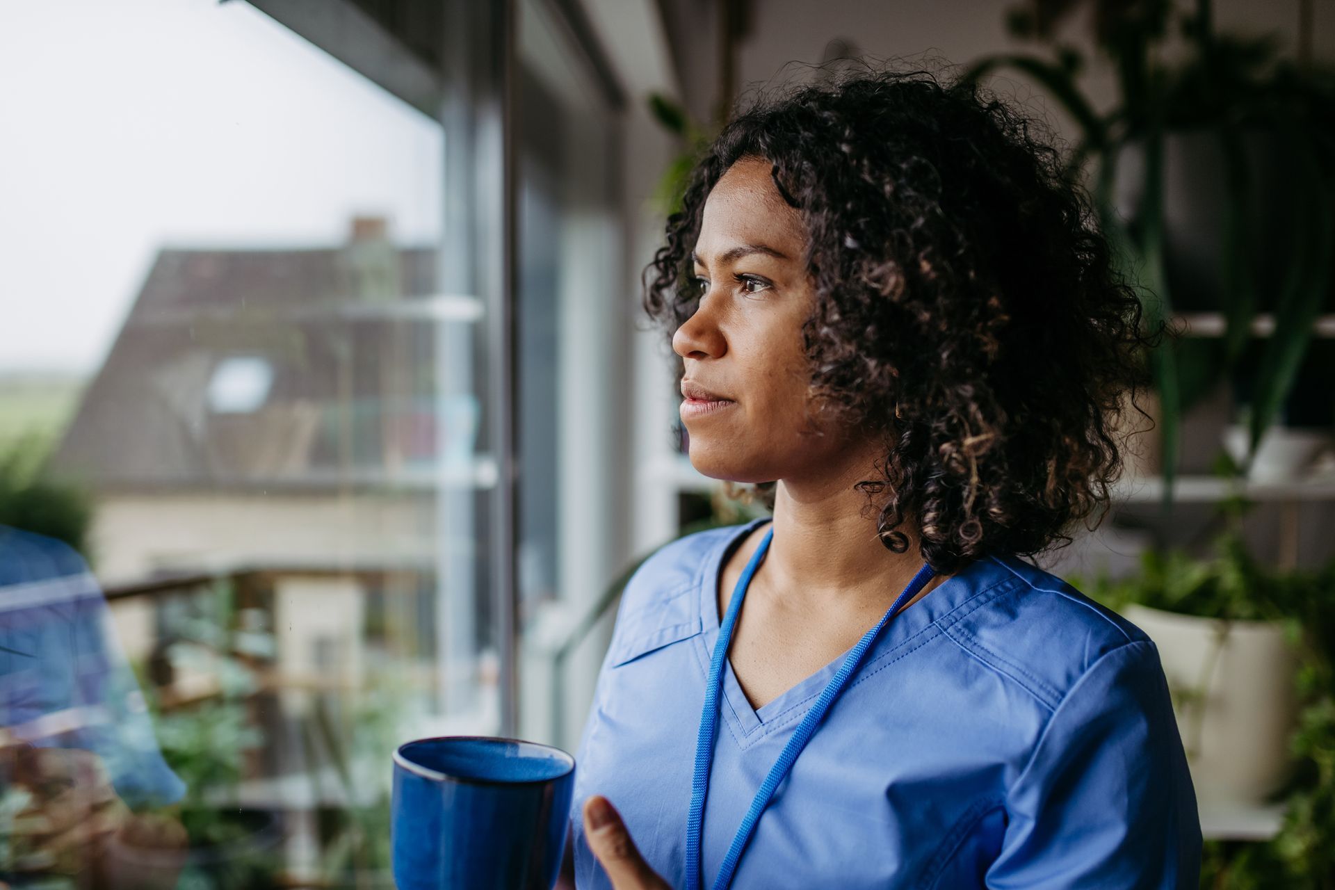 Person in blue scrubs holding a mug and looking out of a window. Person in blue scrubs holding a mug and looking out of a window.
