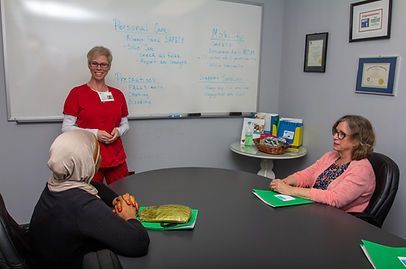 A nurse in red presents to two women seated at a round table. A whiteboard lists notes. A nurse in red presents to two women seated at a round table. A whiteboard lists notes.