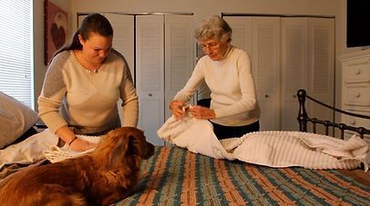Woman and elderly woman folding bedding on a bed with a dog watching in a bedroom. Woman and elderly woman folding bedding on a bed with a dog watching in a bedroom.