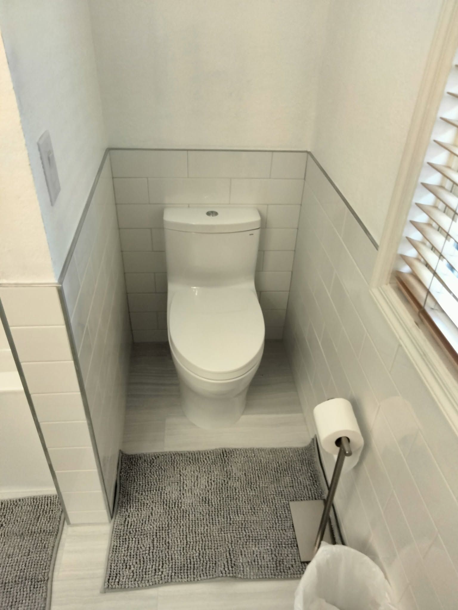 Small white bathroom with a toilet, tiled walls, window blinds, and a toilet brush on the floor.