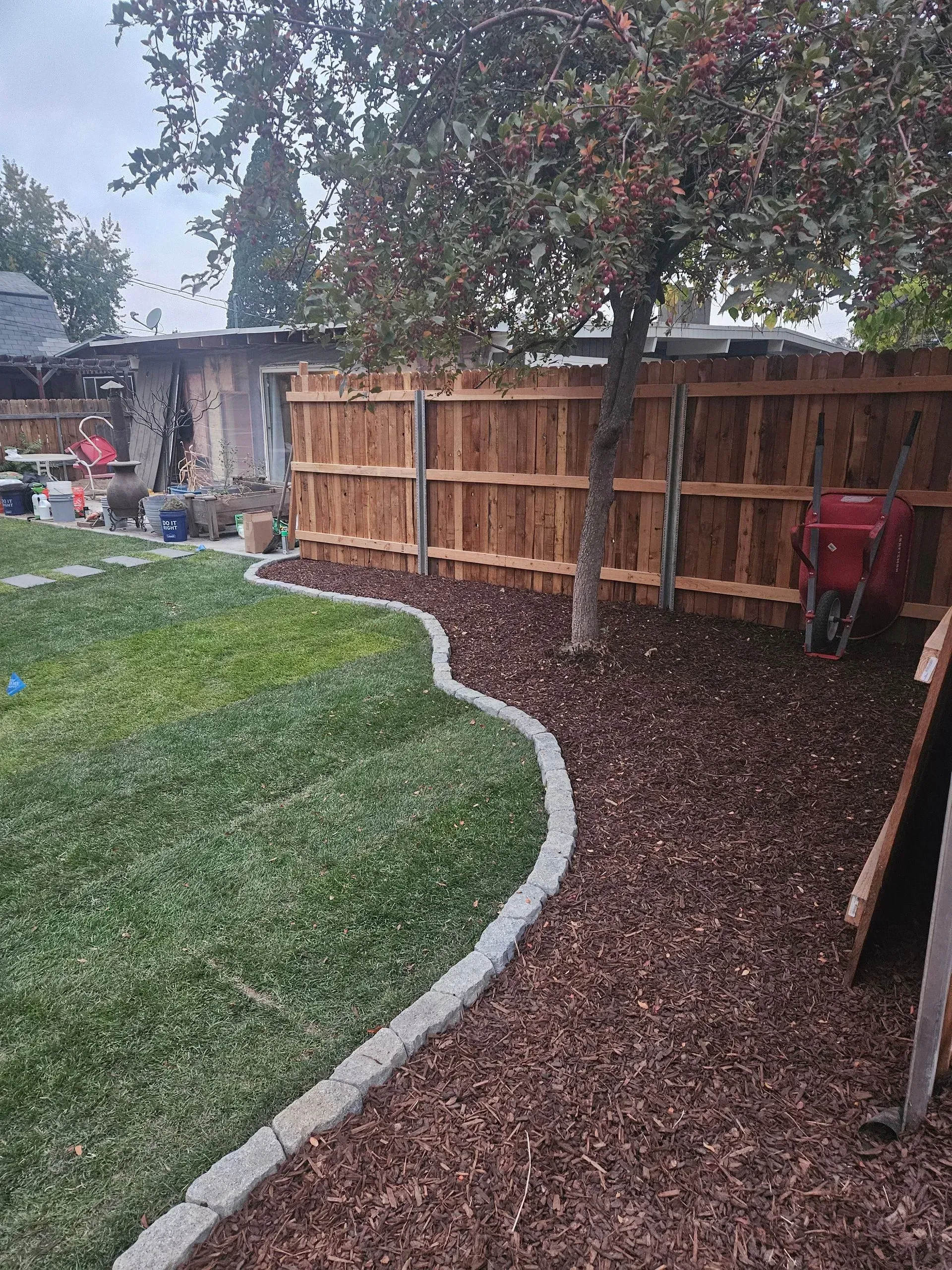 Backyard with curved stone border, green lawn, mulch bed, wooden fence, and tree in light rain