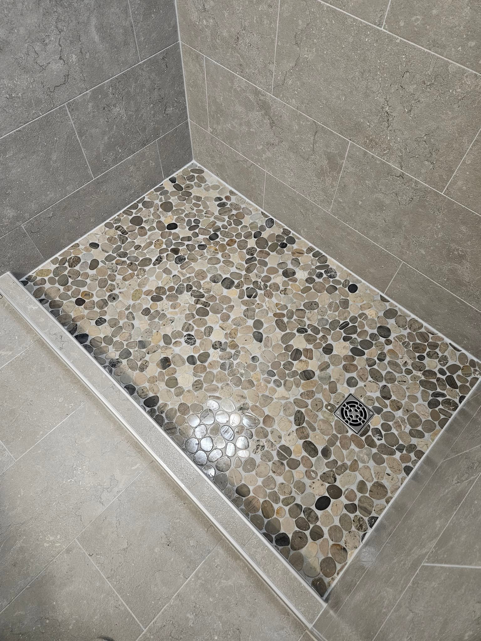 Small tiled shower floor with pebble mosaic base and gray wall tiles, drain visible in corner