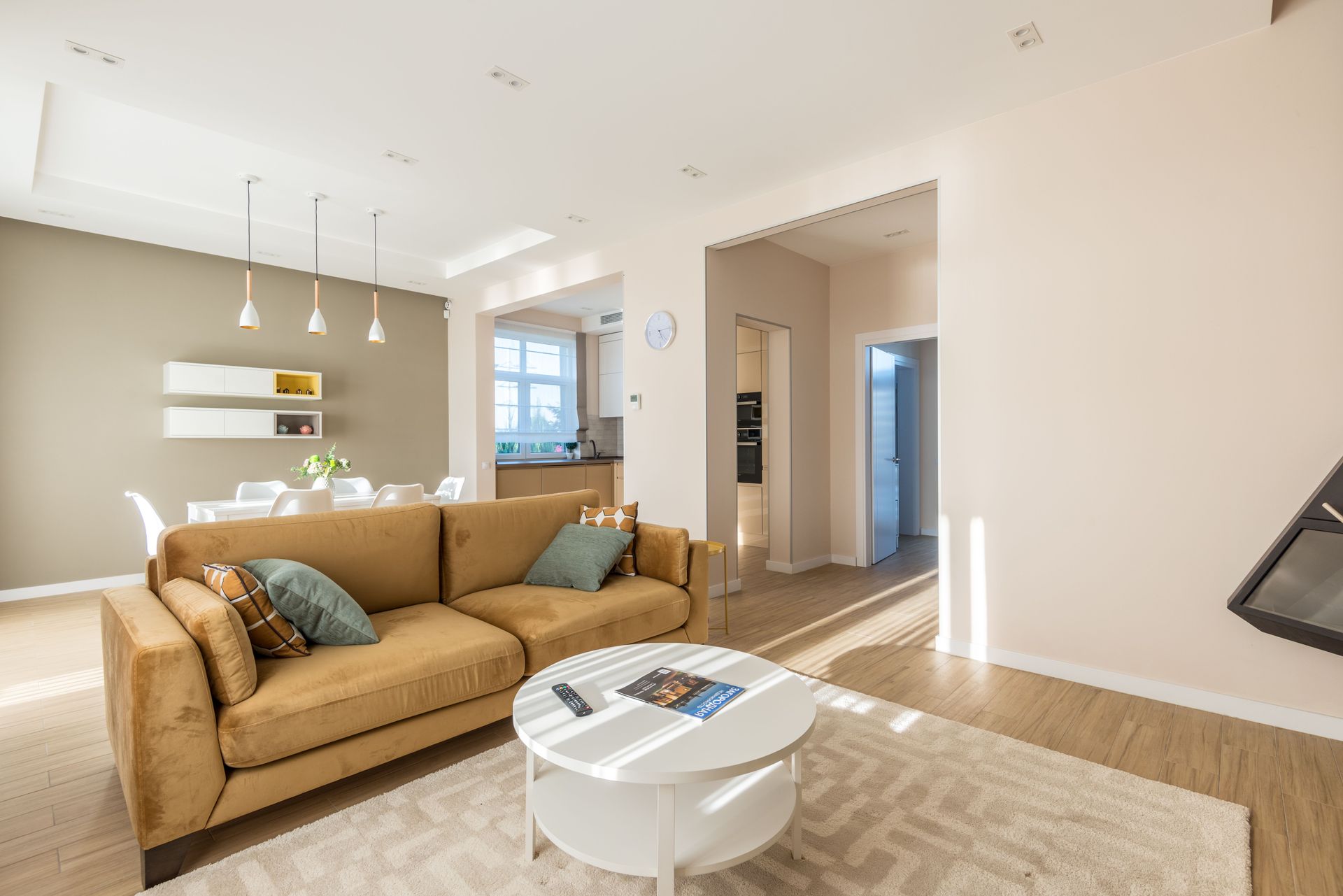 Bright modern living room with tan sofa, round coffee table, rug, and open kitchen in the background