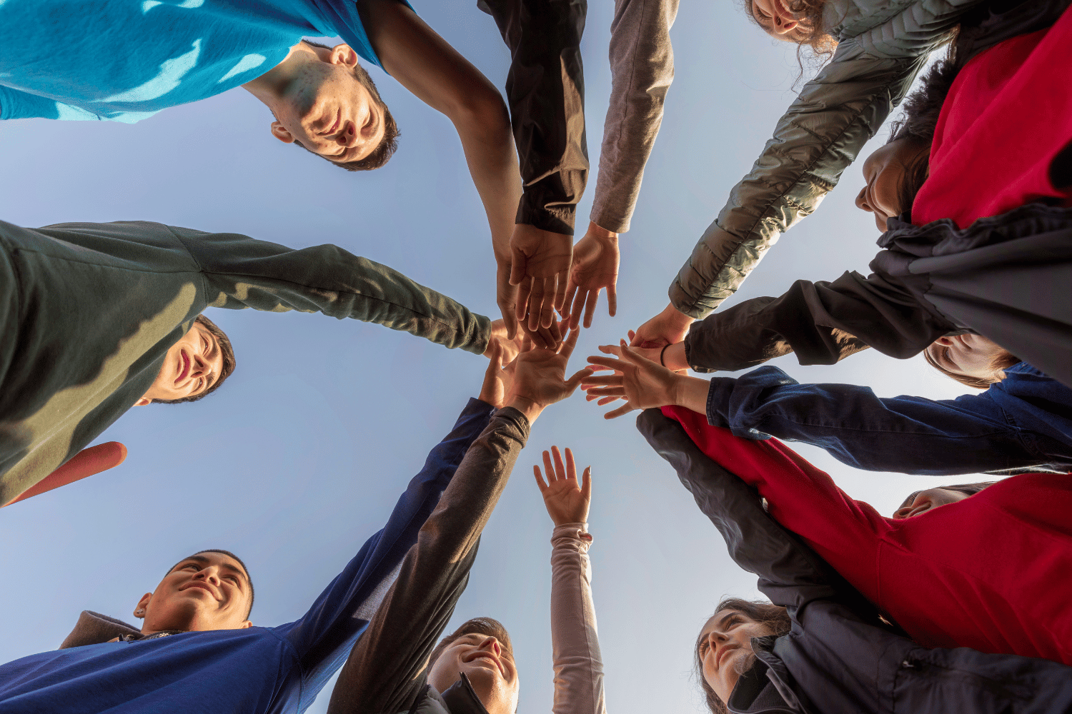 People in a circle stacking hands together against a blue sky, showing teamwork