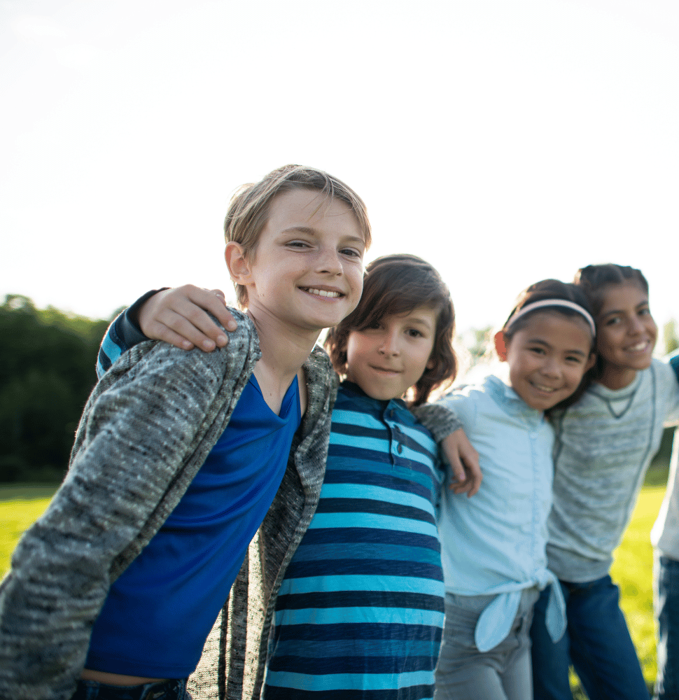 Four smiling children standing arm-in-arm outdoors in a sunny field