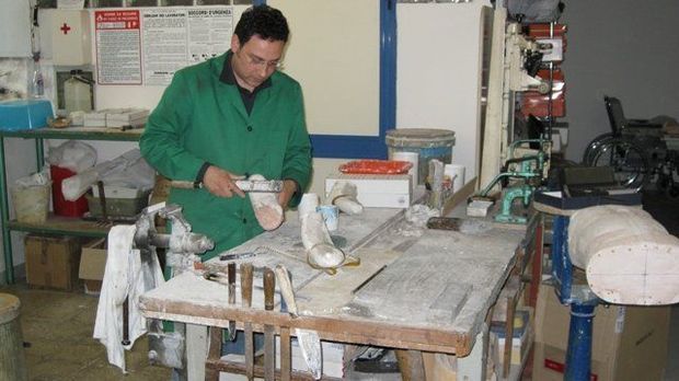 Una persona con una giacca verde lavora a un tavolo da officina ingombro di attrezzi e materiali.