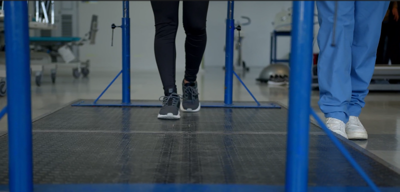 Due persone in piedi sul pavimento di una palestra, dietro delle sbarre blu, con visibili solo le gambe e le scarpe.