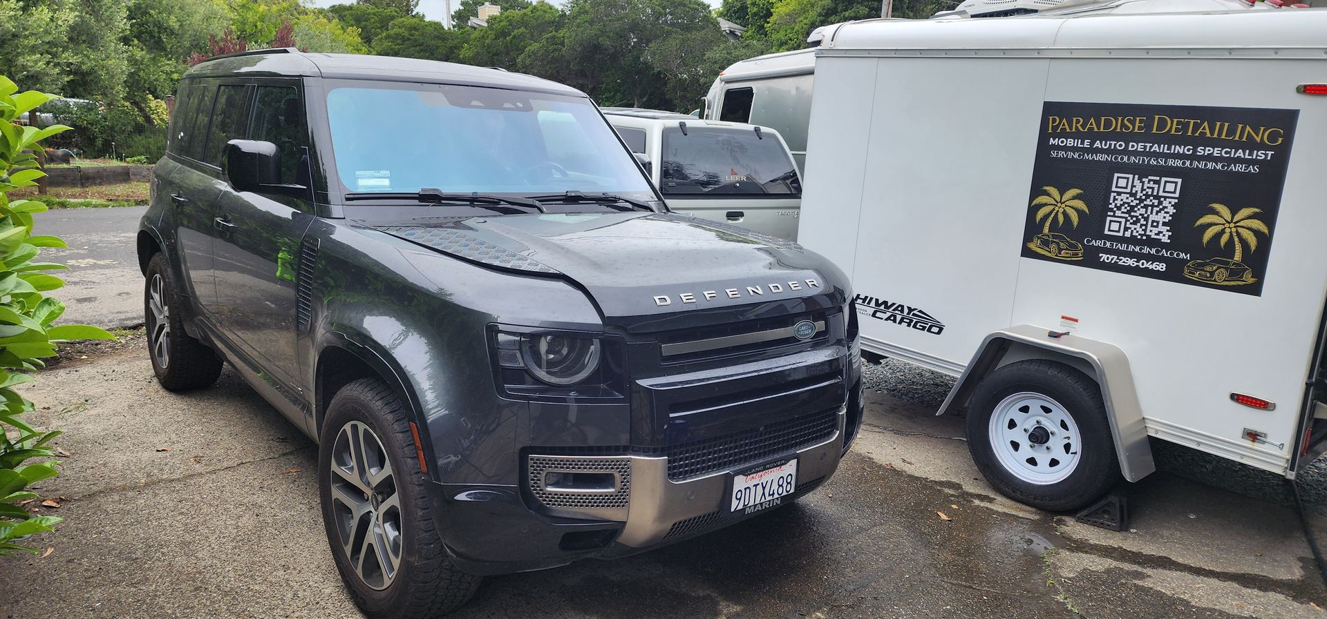 Black SUV parked beside a white trailer in a lot, with other vehicles and trees in the background.