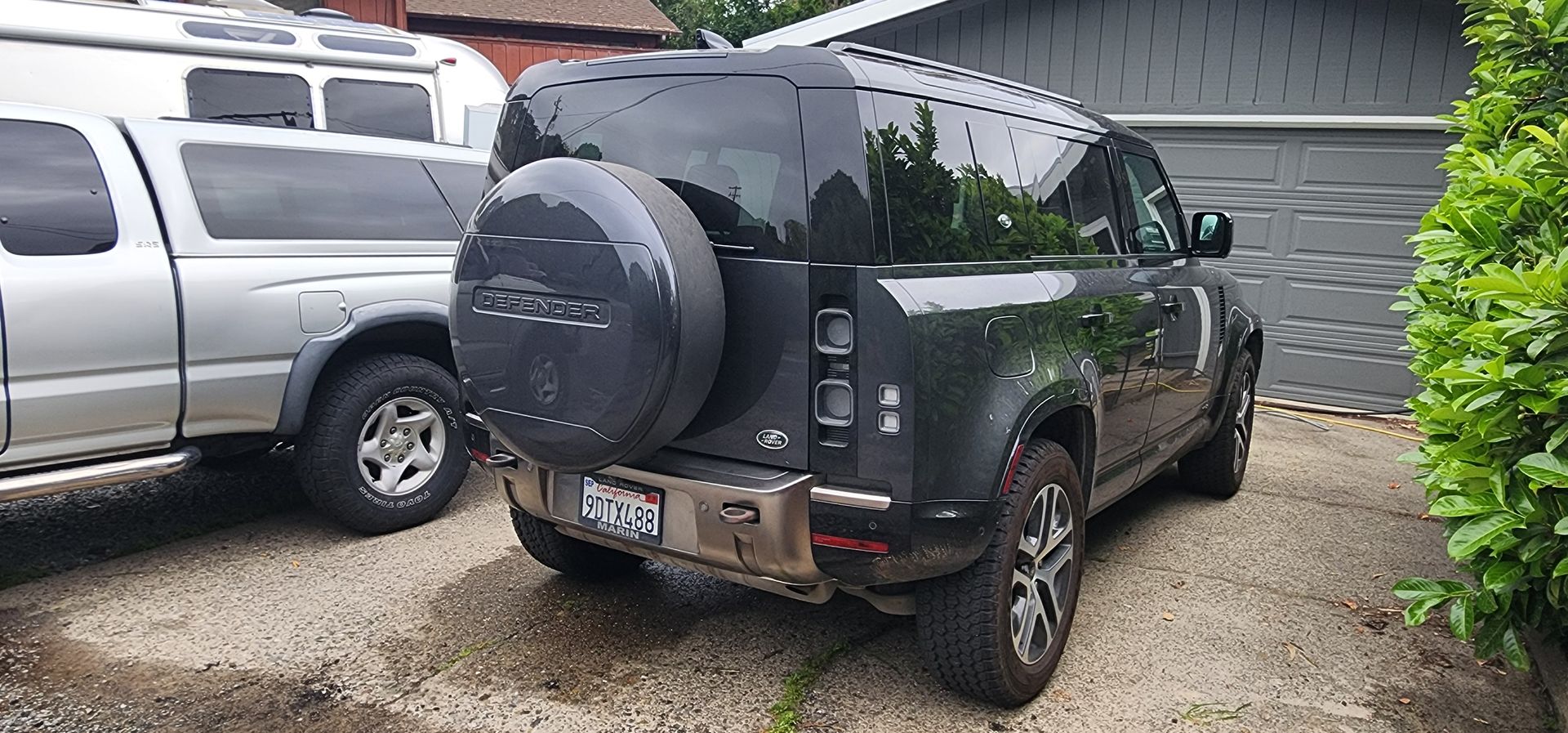 Dark green SUV parked in a driveway beside a silver truck, with a spare tire on the back, in front of a garage