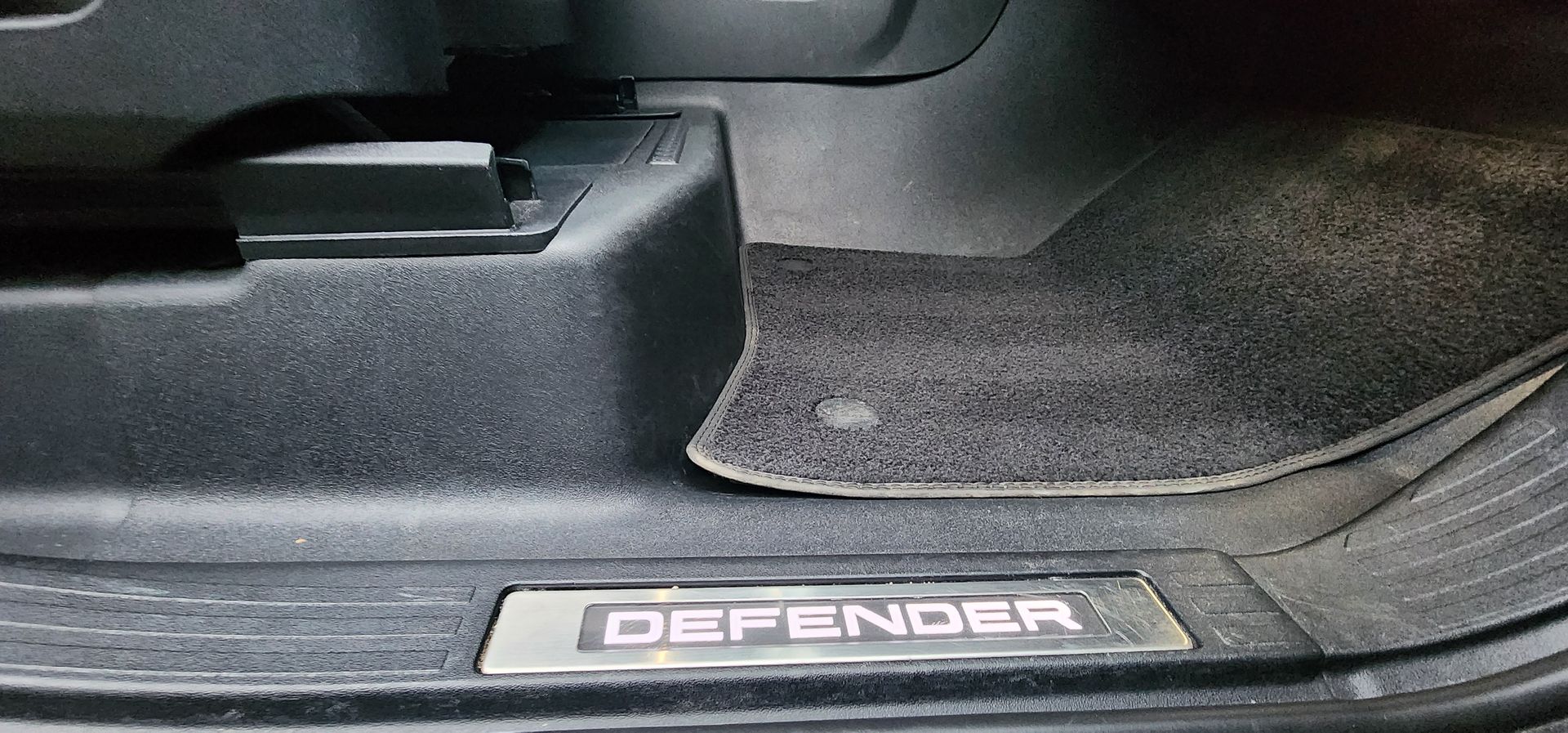 Car door sill with metal DEFENDER badge and black floor mat.