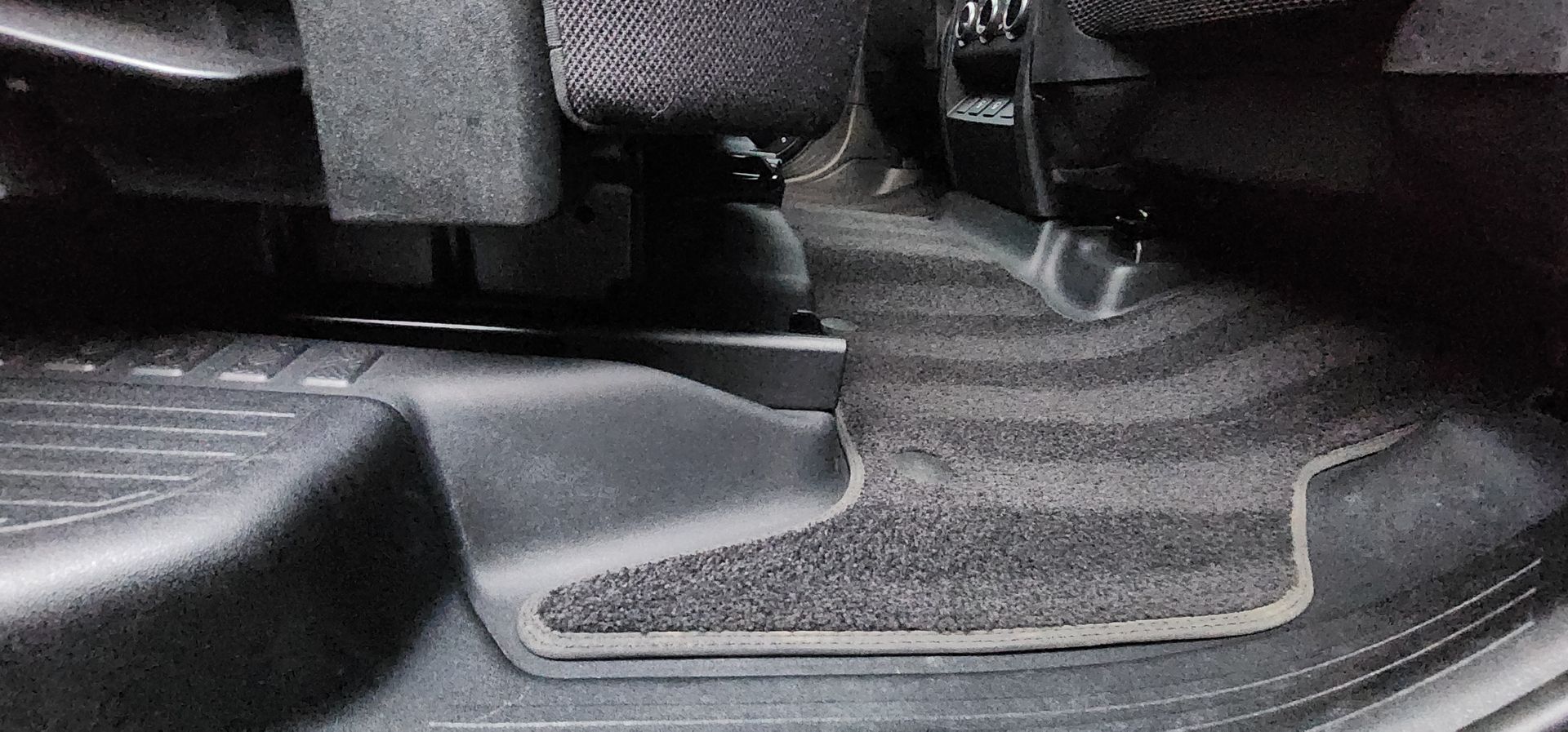 Car floor mat and seat base in a dark vehicle interior, showing textured black rubber flooring.