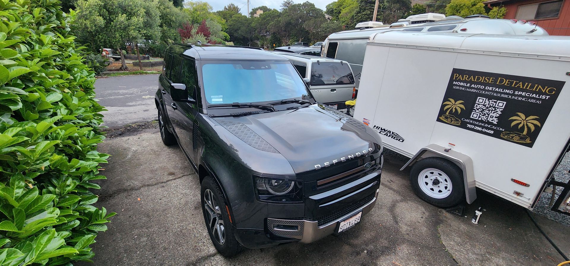 Black SUV parked in a driveway beside a white trailer, with greenery and other cars in the background.
