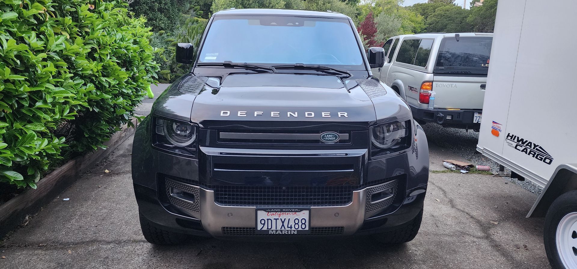 Black Ford Bronco SUV parked in a narrow driveway between a hedge and a white van.