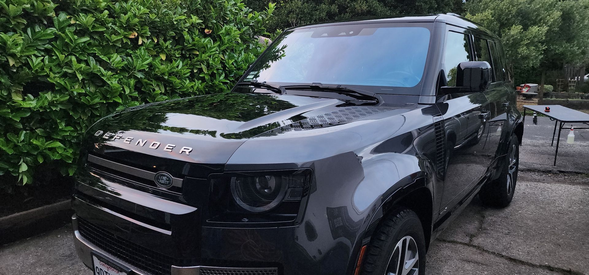 Gray Land Rover SUV parked on a wet driveway beside green bushes and a metal gate.