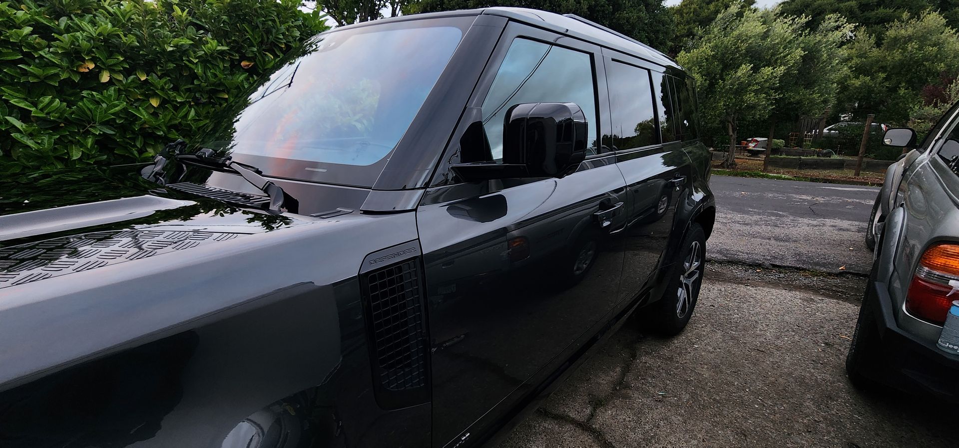 Black vintage car parked outdoors on a wet gravel driveway beside another vehicle, with trees in the background