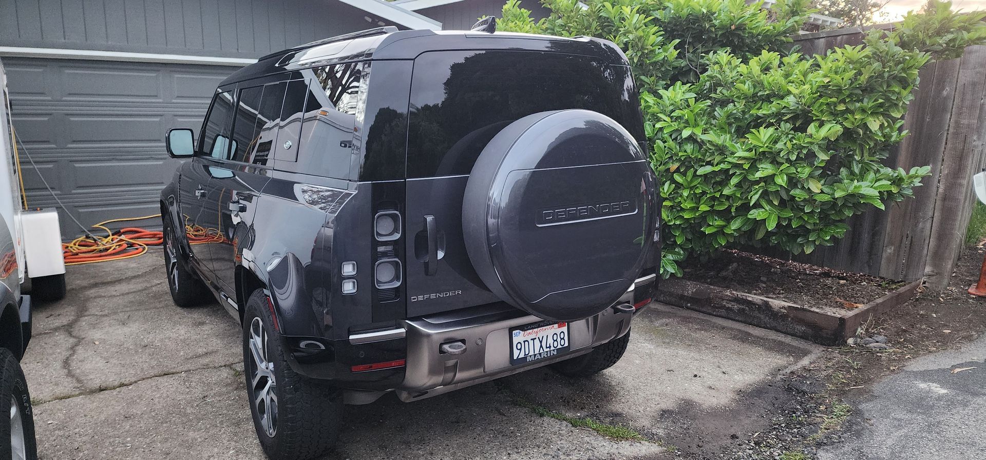Black SUV with rear-mounted spare tire parked in a driveway beside a garage and hedge