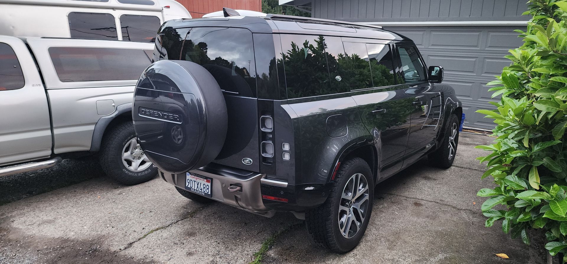 Black SUV parked in a driveway beside a silver vehicle and a green hedge, wet from rain